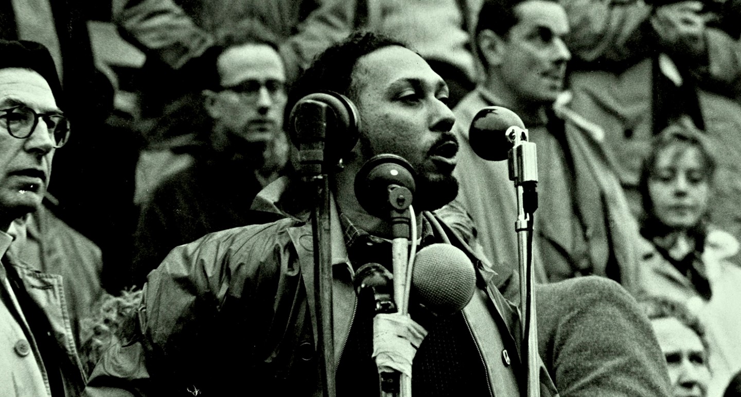 Black and white photograph: Stuart Hall depicted behind several microphones and addressing a crowd (who are presumably located behind the camera). A variety of people (mostly white and wearing coats) are visible in the background.